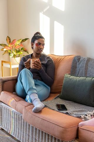 Sunlit indian woman sipping tea on wicker sofa with under-eye patches for cozy self-care