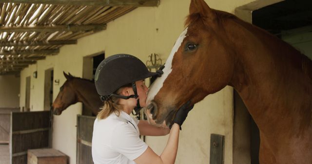 Woman in equestrian gear gently interacts with horse in stable. Ideal for themes of animal care, human-animal bond, equestrian activities, rural life, and farm settings.