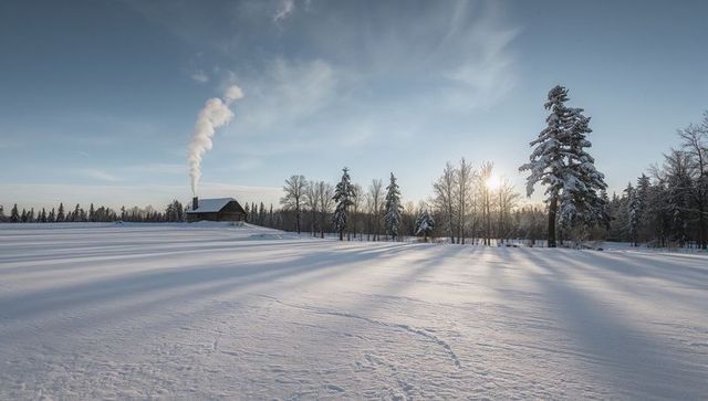 Snow-covered farmhouse sitting on rise sending chimney smoke across sunlit winter field