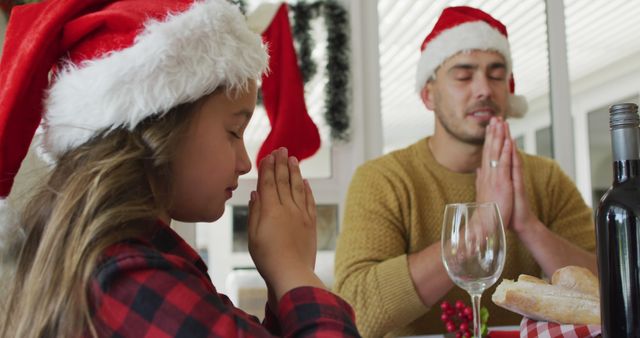 Father and daughter, both wearing Santa hats, spending quality time together praying at a dining table during a Christmas celebration. The table is adorned with festive decorations including a bottle of wine, glasses and bread. Use this image for promoting family-friendly holiday gatherings, festive celebration events, or content focusing on Christmas traditions and family bonding.