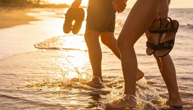 Couple walking barefoot at sunset holding sandals on golden beach with sand reflections