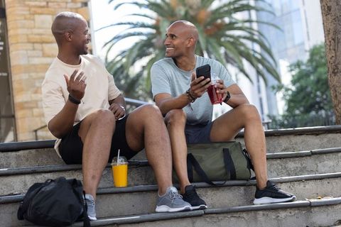 Two african american men chatting and laughing on outdoor steps with drinks and smartphone