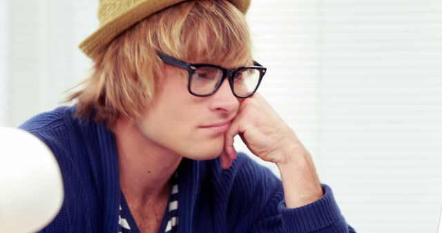 Depicts a young man wearing glasses and a hat, sitting indoors and appearing thoughtful. He is dressed casually, creating a relaxed and modern vibe. Ideal for content related to modern lifestyle, study, work-at-home scenarios, creative thinking, millennials. Useful for blogs, articles, advertisements, or websites focusing on youth, fashion, and contemporary culture.