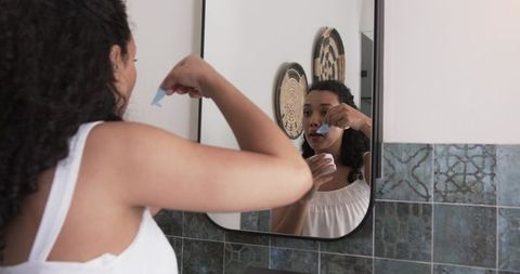 African woman applying blue pore strip in bathroom mirror during morning skincare routine
