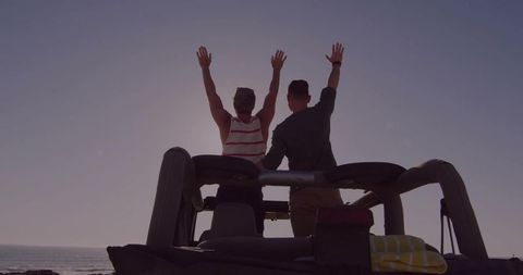 Two Friends Raising Arms on Open-Top Jeep Overlooking Ocean Sunset, Coastal Road Trip