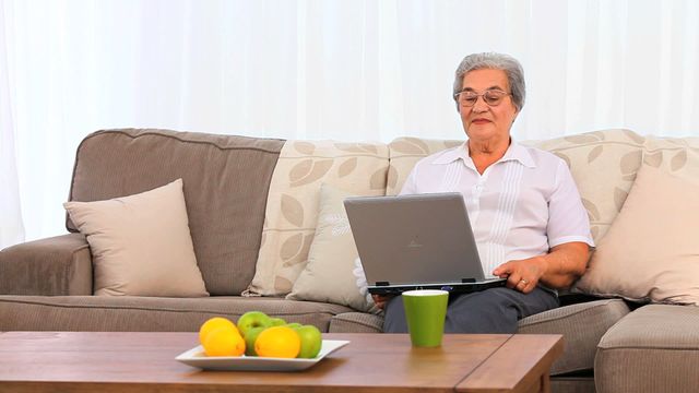 Senior woman sitting on a comfortable couch, working on a laptop with a focused expression. A green coffee cup and a bowl of fresh fruits on a nearby wooden table suggest a relaxed and homey environment. This video can be used to depict concepts such as remote work, elderly people using technology, relaxation at home, and a modern, comfortable lifestyle for seniors.