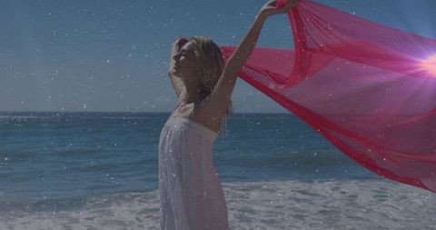 Woman embracing breeze on sunlit beach while holding flowing pink scarf and sea silhouette