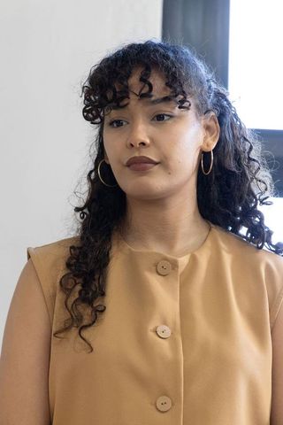 Curly-haired professional woman wearing tan buttoned top and gold hoops, backlit headshot