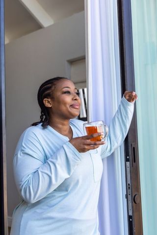 African american woman enjoying morning tea at sliding door in light blue loungewear