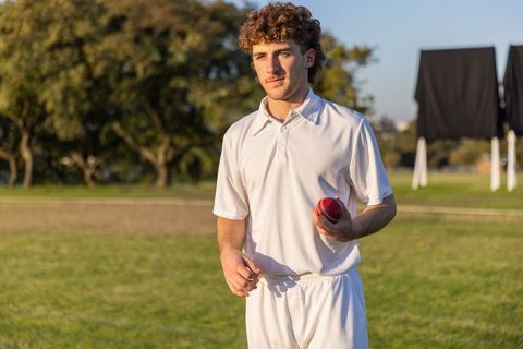 Young cricketer holding red ball on sunlit field wearing white uniform during practice