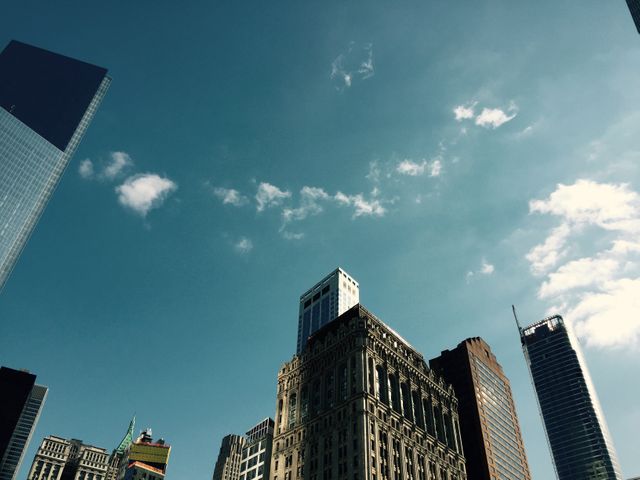 Photo of urban skyscrapers with clear blue sky and a few clouds in the background. Tall buildings showcasing different architectural styles. Ideal for use in projects related to urban development, real estate, city planning, architecture designs, metropolitan travel brochures, and skyline enthusiasts.