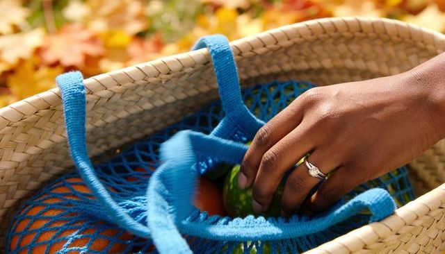 Right hand reaching green fruit in blue mesh tote inside straw basket with autumn leaves