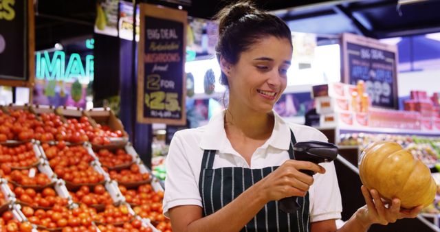 A young female employee wearing an apron is scanning a pumpkin in the produce section of a grocery store. The background features an abundant display of fresh vegetables and vibrant signs. This stock photo is ideal for illustrating themes related to retail work, customer service, in-store grocery shopping, and healthy eating. It can be used in articles, blogs, promotional materials, and websites that focus on food, commerce, employee training, and supermarket settings.