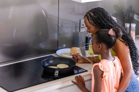 African american mother and daughter cooking pancakes together in modern kitchen, smiling