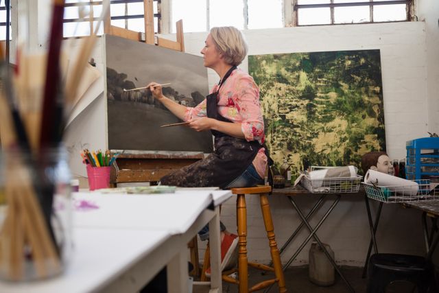 Woman painting on canvas in an art studio workshop. Featured is an artist actively working on her project, surrounded by art supplies like brushes and paints. Suitable for use in articles or promotions related to artistic expression, creative workshops, and art education.