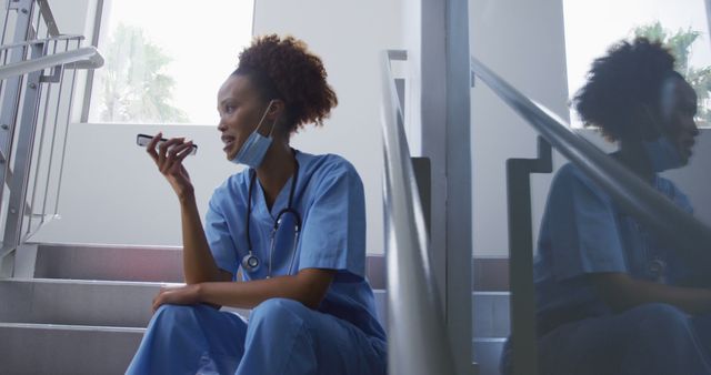 Biracial female doctor wearing face mask sitting on stairs in hospital talking on smartphone - Download Free Stock Photos Pikwizard.com