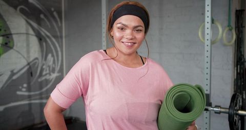 Smiling woman holding green yoga mat in industrial gym, wearing pink top and headband