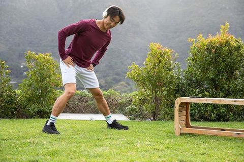 Asian man stretching outdoors with wireless headphones on green lawn near wooden bench