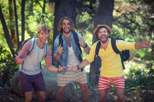 Three young men enjoying an outdoor adventure in a forest on a sunny day. They are smiling and posing for the camera, wearing casual summer clothing and backpacks. Perfect for promoting outdoor activities, travel, friendship, and leisure time.