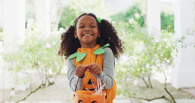 Young girl dressed in a pumpkin costume during Halloween, happily trick or treating outdoors. Her curly hair and joyful smile create a sense of fun and celebration. Great for promotions related to Halloween events, children's costumes, outdoor activities, and festive advertising.