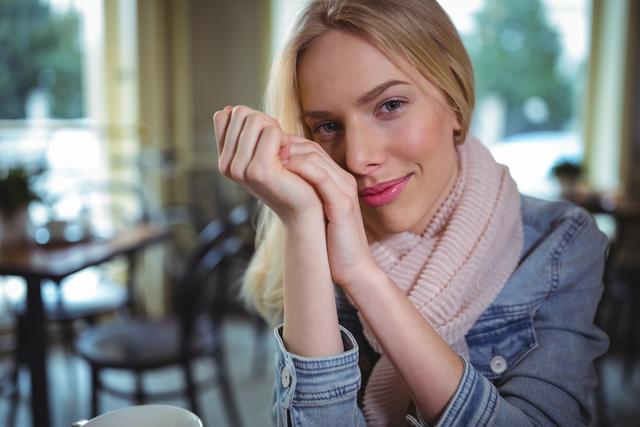 Woman sitting in a cozy cafe, smiling warmly while wearing a denim jacket and scarf. Ideal for use in lifestyle blogs, advertisements for cafes or coffee shops, and content promoting relaxation and leisure.
