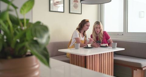 Young women sharing smartphone and cake at cozy kitchen banquette, casual coffee break