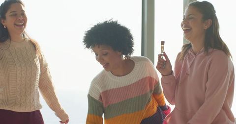 Diverse friends laughing by window in colorful knitwear holding makeup brush