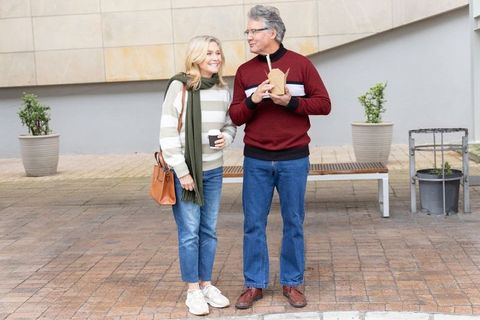 Senior couple sharing coffee and takeout while strolling urban plaza in cozy sweaters
