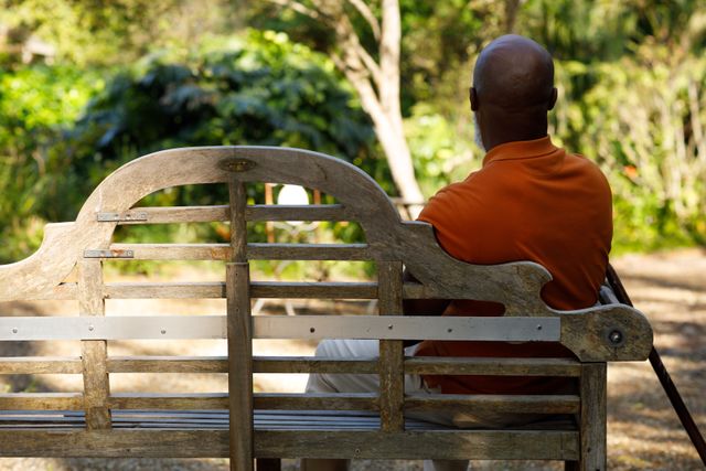 Rearview of an old african-american man sitting on a wooden park bench with his cane. he's wearing an orange shirt with cream pants