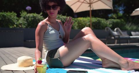 African american woman relaxing poolside with cocktail, sun hat and smartphone on towel
