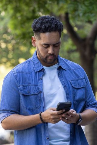 African american man checking smartphone in park wearing denim shirt and beaded bracelet