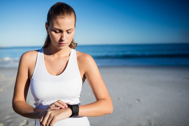 Young woman in sportswear using smart watch on beach. Ideal for promoting fitness technology, outdoor exercise, healthy lifestyle, and summer activities. Suitable for advertisements, blogs, and articles related to fitness tracking, wellness, and beach workouts.
