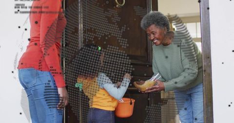 Senior woman handing out halloween candy to child at doorway with parent present