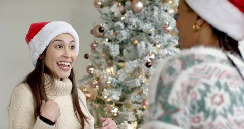 Diverse friends laughing and chatting by frosted Christmas tree wearing Santa hats, cozy knitwear