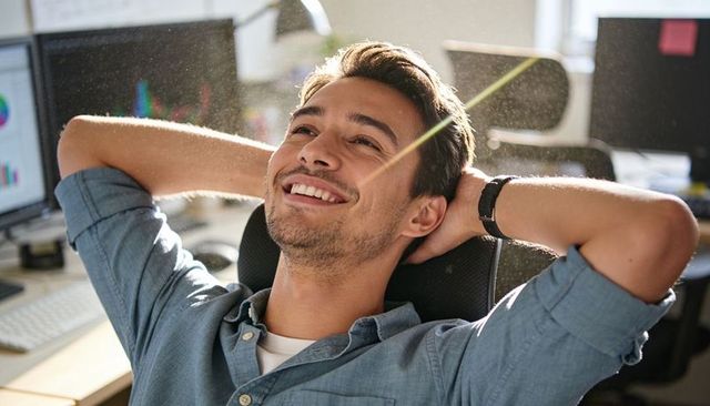 Young professional relaxing at desk leaning back smiling basking in sunlit modern office