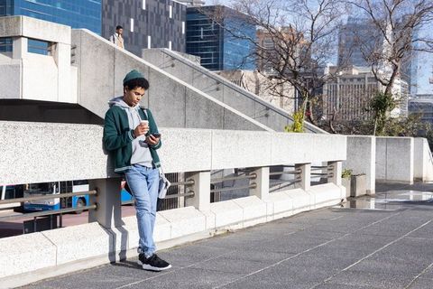 African american commuter leaning on urban railing, checking phone and holding coffee