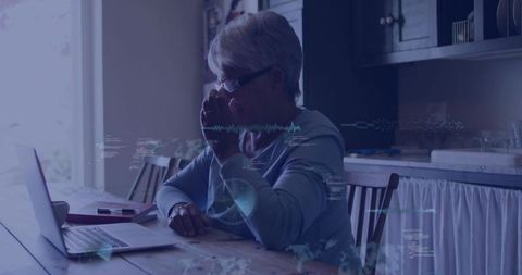 Senior woman using laptop at sunlit kitchen table with digital data overlays for telehealth