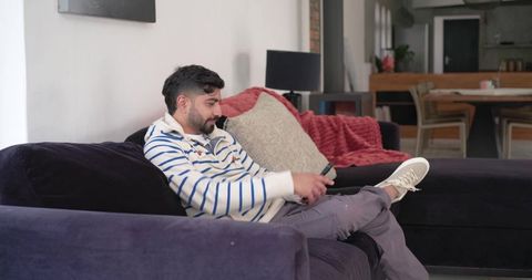 Indian man relaxing on purple sectional sofa holding remote in cozy open-plan living area