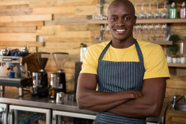 Smiling male barista standing with arms crossed in a coffee shop, wearing a striped apron and yellow shirt. Coffee machines and glasses are visible in the background. Ideal for use in articles or advertisements related to coffee shops, barista training, customer service, small businesses, and hospitality industry.