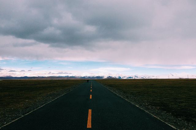This image features an empty road stretching towards the horizon, flanked by an expansive landscape under a cloudy sky. The image evokes a sense of solitude and adventure, making it ideal for travel blogs, scenic journey promotions, and motivational posters about journeys or reaching goals.