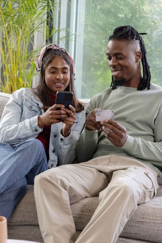 Diverse couple using mobile payment on smartphone while sitting on sofa with credit card