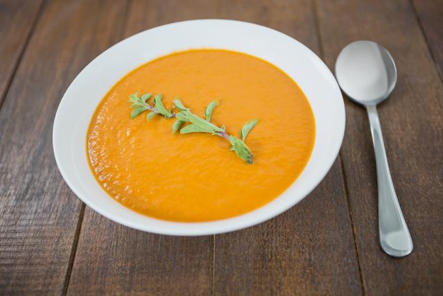 Bowl of creamy pumpkin soup garnished with fresh herbs, placed on a rustic wooden table with a spoon beside it. Ideal for use in food blogs, recipe websites, healthy eating promotions, and autumn-themed content.