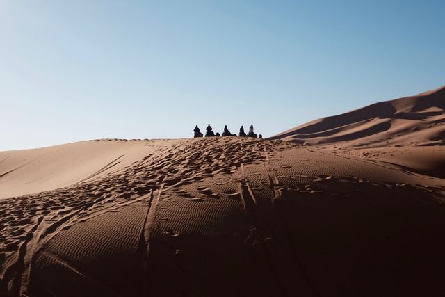 Group of travelers silhouetted against setting sun, sitting atop a sand dune in vast desert, creating a scene of tranquility and grandeur. Ideal for promoting travel, adventure, outdoor activities, and tourism campaigns. Effective in illustrating concepts like exploration, solitude, and the beauty of nature.