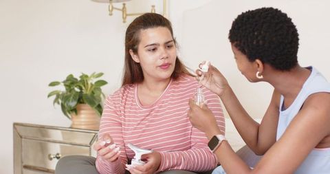 Diverse friends testing skincare serum dropper and applicator on cozy living room couch