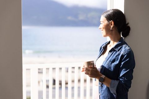 Woman enjoying morning coffee on ocean-view balcony, checking smartwatch, coastal lifestyle