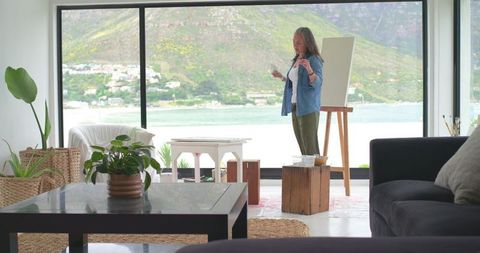 Mature woman painting on canvas in sunlit coastal living-room studio with mountain view
