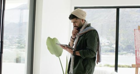 Contemplative south asian man holding tablet in minimalist sunlit lakeside creative studio