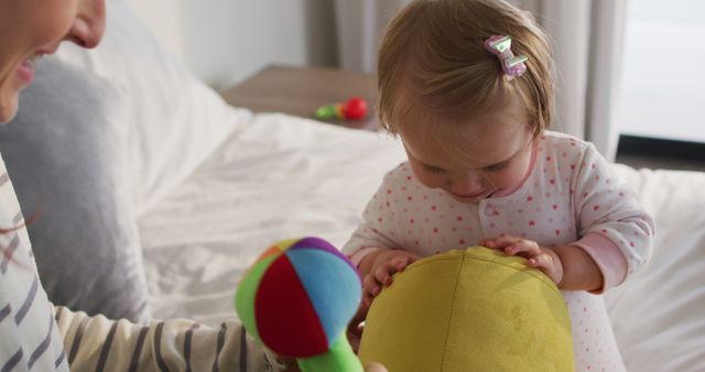 Mother and baby bonding while playing with colorful toys on bed. Baby holding a green soft ball with curiosity, while mother enjoys the moment. Ideal for family-themed content, parenting blogs, baby product advertisements, and articles on early childhood development.