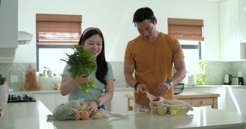 Asian couple unpacking groceries preparing fresh produce on sunlit modern kitchen island