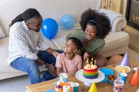 Black family celebrating child's birthday at home with rainbow cake, balloons, joyful smiles